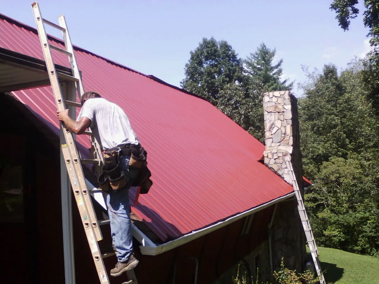Roofer on a ladder beside a red metal roof and stone chimney — Soffit & Fascia Repair by Roofing Brothers in Montclair